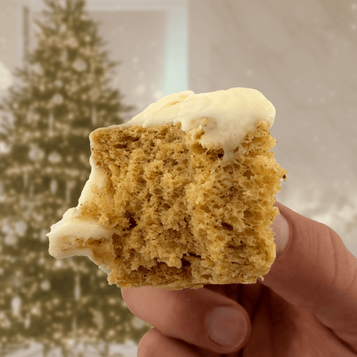 Gingerbread Protein Cake Mix with creamy frosting held in hand, holiday tree in background, cozy winter vibes.