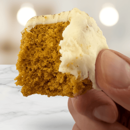 Close-up of a hand holding a moist slice of Pumpkin Spice Protein Cake Mix topped with creamy frosting.